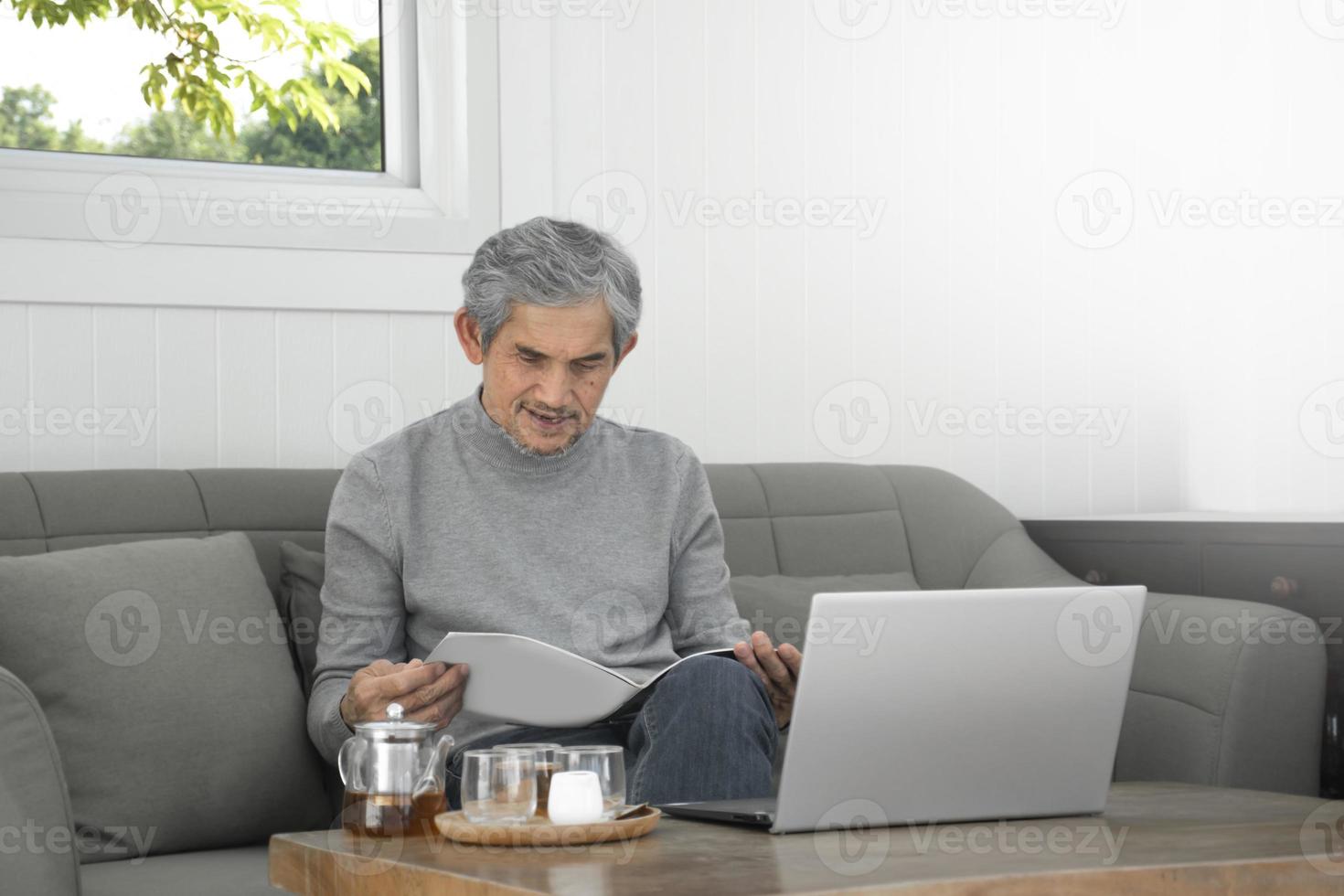 Portrait d'un homme asiatique senior âgé assis près d'une fenêtre en verre le matin pour travailler à domicile et vérifier ses affaires sur son ordinateur portable sur la table avec une mise au point sérieuse, douce et sélective. photo