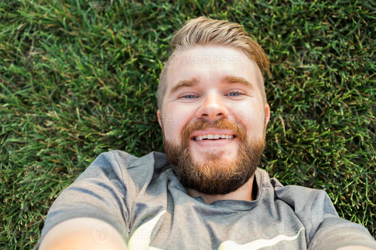 jeune homme avec barbe et style de cheveux de mode allongé sur l'herbe prenant selfie - tenant un smartphone ou une tablette et regardant la caméra. photo