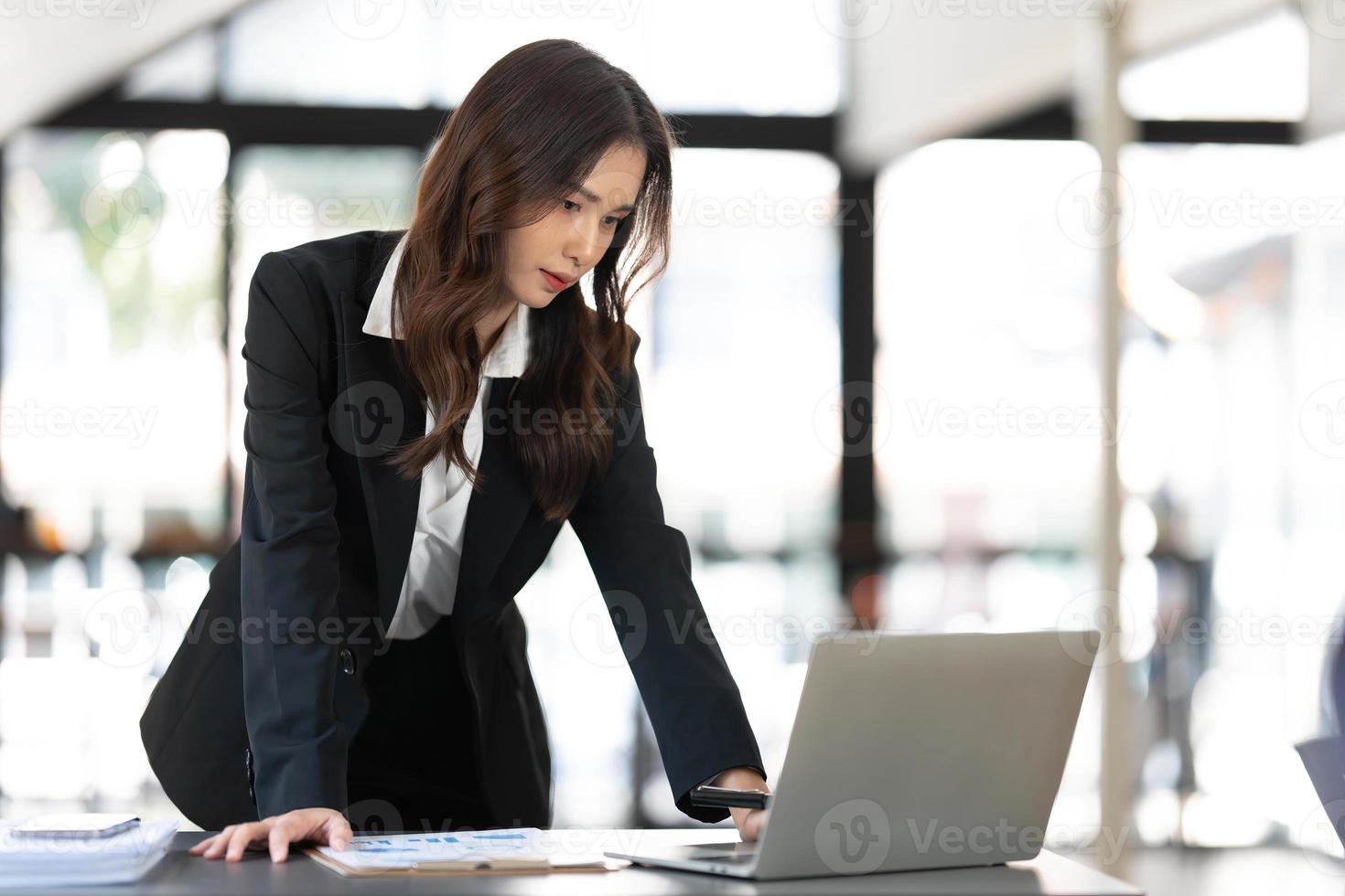 jeune femme d'affaires concentrée debout à table au bureau, utilisant un ordinateur portable, regardant un écran d'ordinateur, lisant ou écrivant des e-mails professionnels, recherchant des informations sur Internet, travaillant sur un projet. photo