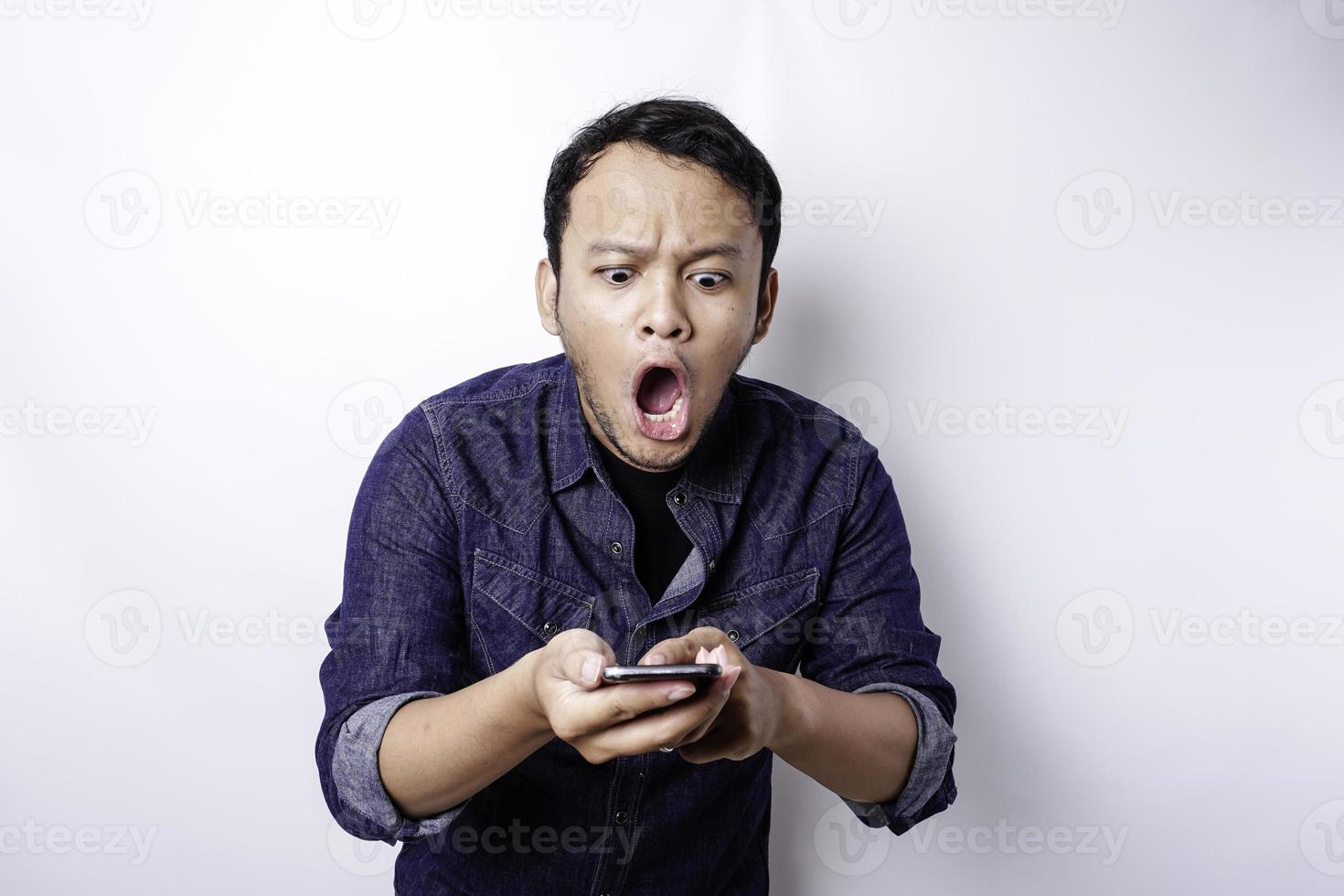 homme asiatique choqué portant une chemise bleue et tenant son téléphone, isolé sur fond blanc photo