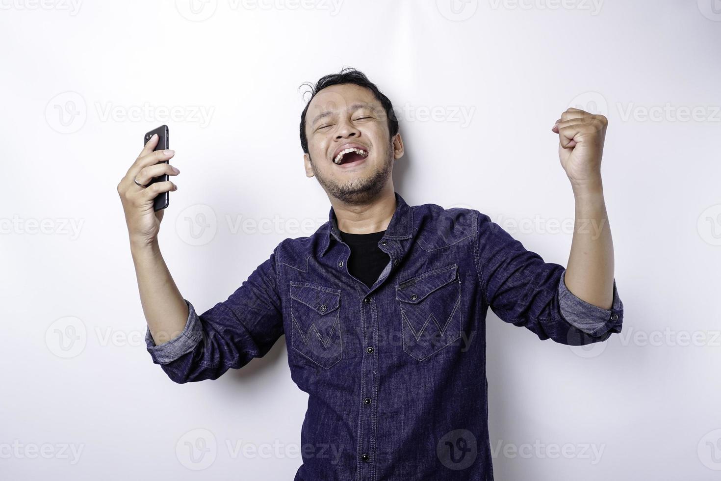 un jeune homme asiatique avec une expression heureuse et réussie portant une chemise bleue et tenant son téléphone, isolé sur fond blanc photo