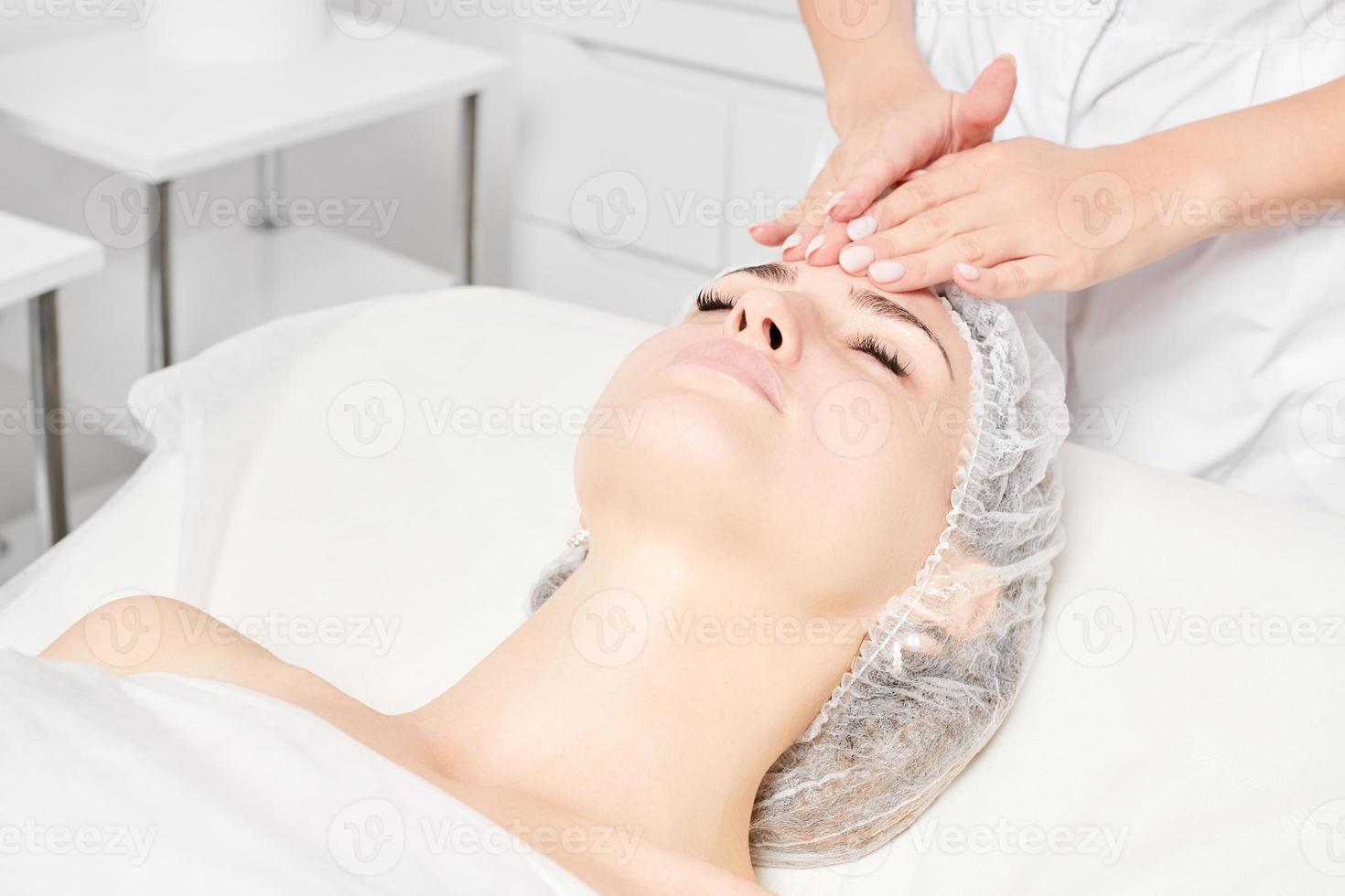 le cosmétologue masse le masque crème dans la peau du visage de la femme pour le rajeunissement, procédure dans un salon de beauté photo