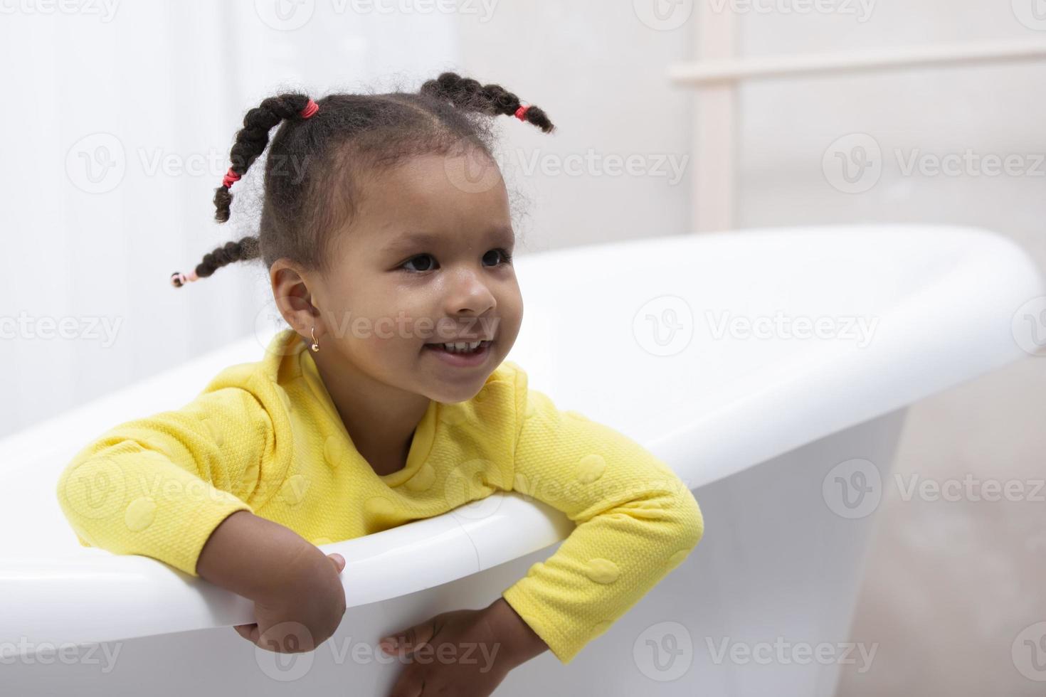 petite fille afro-américaine vêtue d'une robe jaune avec des nattes bouclées est assise dans un bain rétro. photo