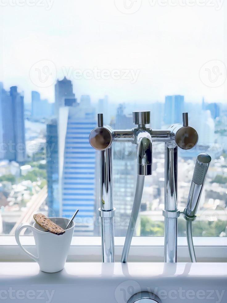 salle de bain image de style coréen, baignoire en céramique blanche de luxe moderne, ensemble de robinets en chrome poli, une tasse de café blanche avec biscotti sur le dessus, avec vue sur la ville floue en arrière-plan photo