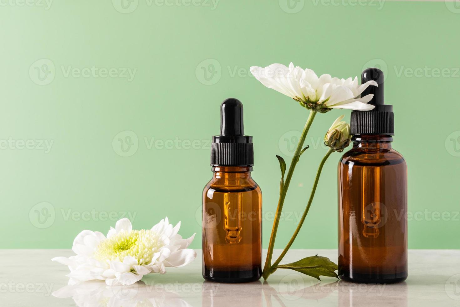 vue de face de deux flacons cosmétiques sans nom avec un compte-gouttes en verre foncé avec des chrysanthèmes blancs sur fond vert et un podium en marbre. maquette. photo