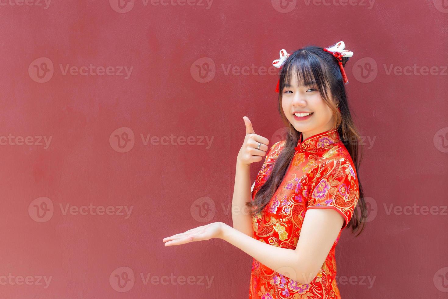 jolie fille asiatique aux cheveux longs qui porte une robe cheongsam rouge sur le thème du nouvel an chinois alors qu'elle montre sa main pour présenter quelque chose sur fond rouge. photo