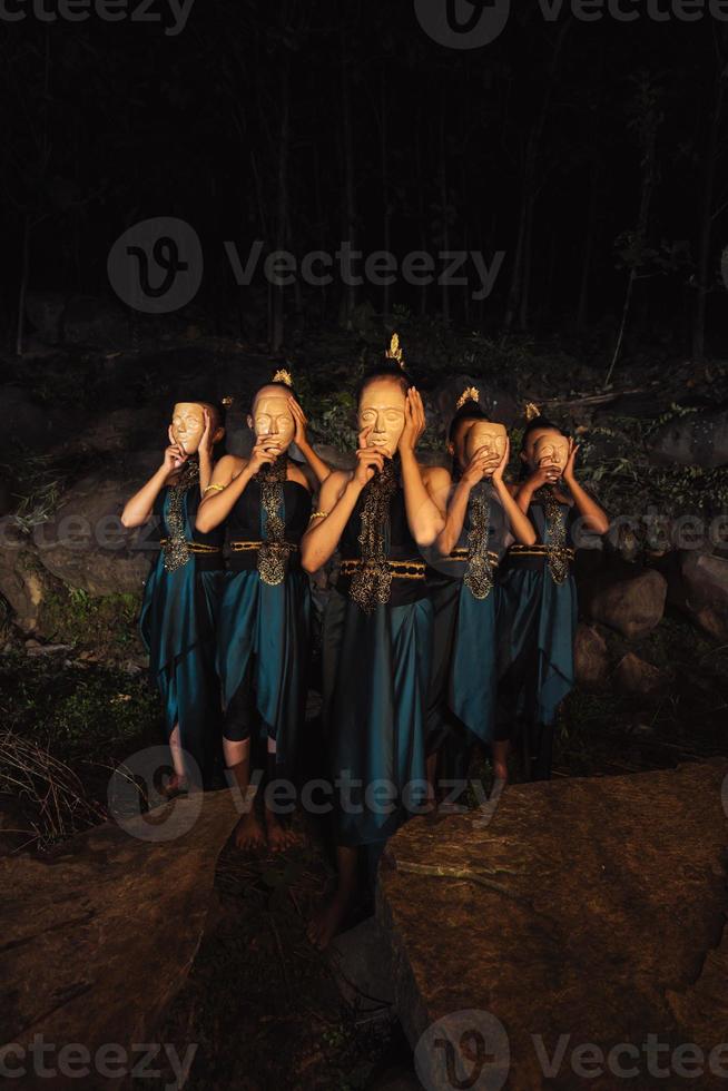 un groupe de femmes asiatiques portant des masques en bois en costumes verts tout en se tenant entre les rochers à l'intérieur de la forêt photo