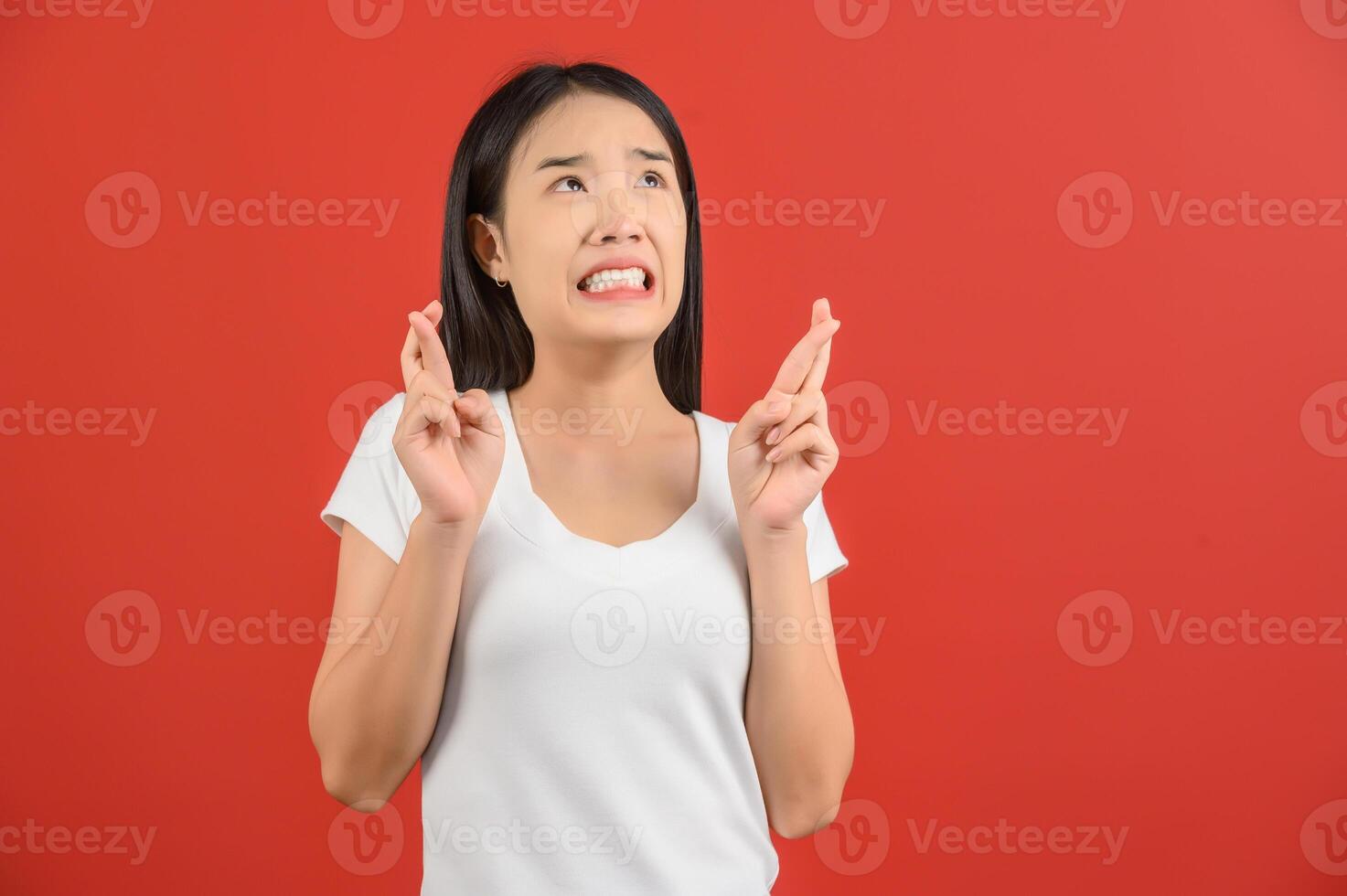 portrait d'une jeune femme asiatique inquiète en t-shirt blanc tenant les doigts croisés pour la bonne chance, exprimant un grand désir de gagner isolé sur fond rouge photo