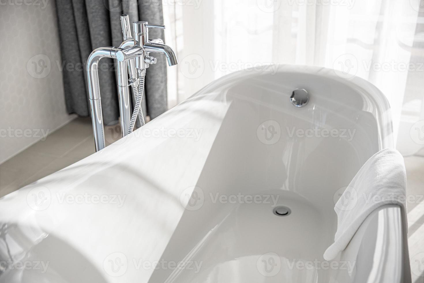 robinets chromés et mitigeur sur baignoire en céramique blanche dans une salle de bain blanche de style rétro dans une maison de luxe à proximité d'un rideau blanc sous la lumière du soleil. intérieur de salle de bain idyllique photo