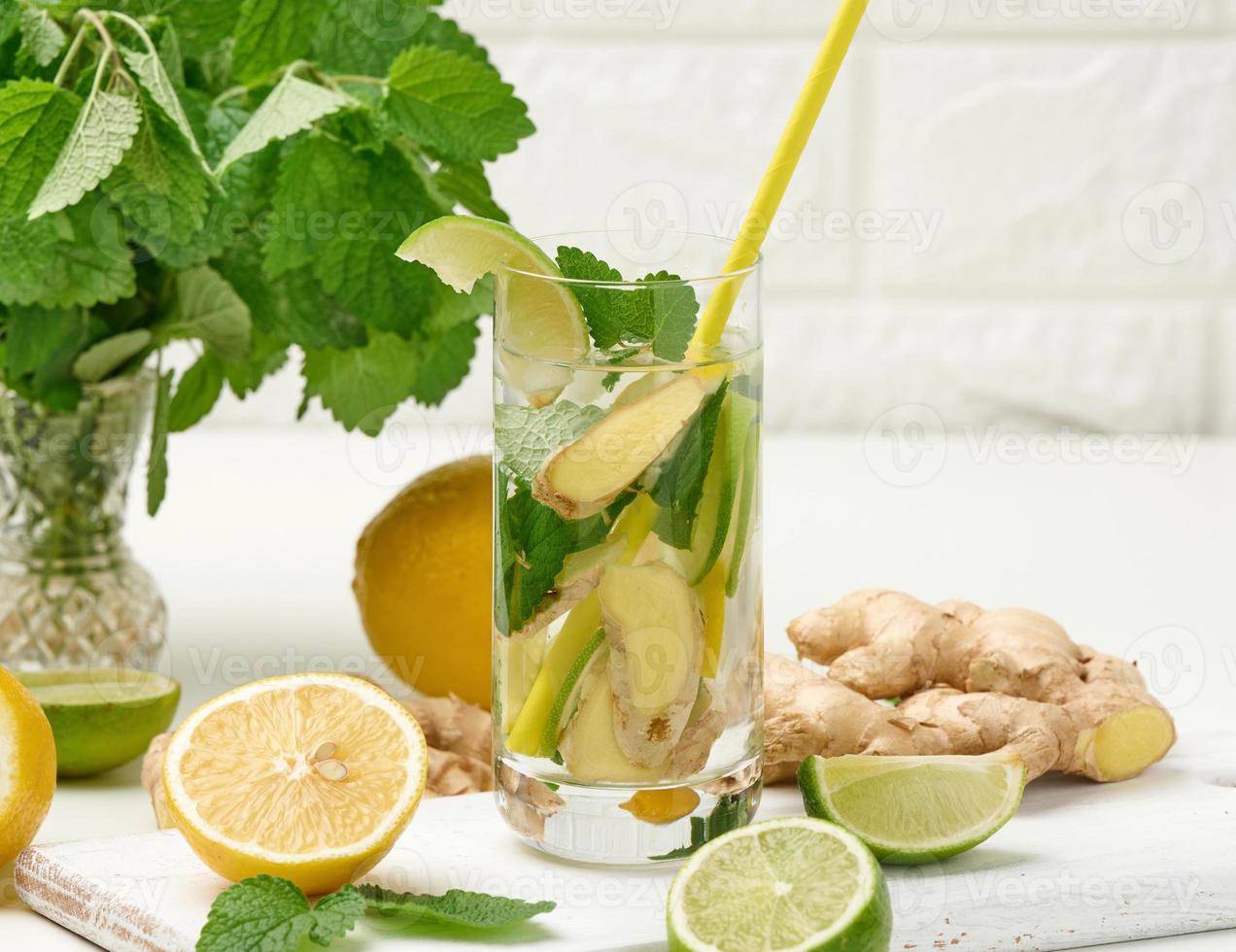 tasse en verre transparent avec citron, morceaux de citron vert et feuilles de menthe et morceaux de gingembre sur un tableau blanc, désintoxication. photo