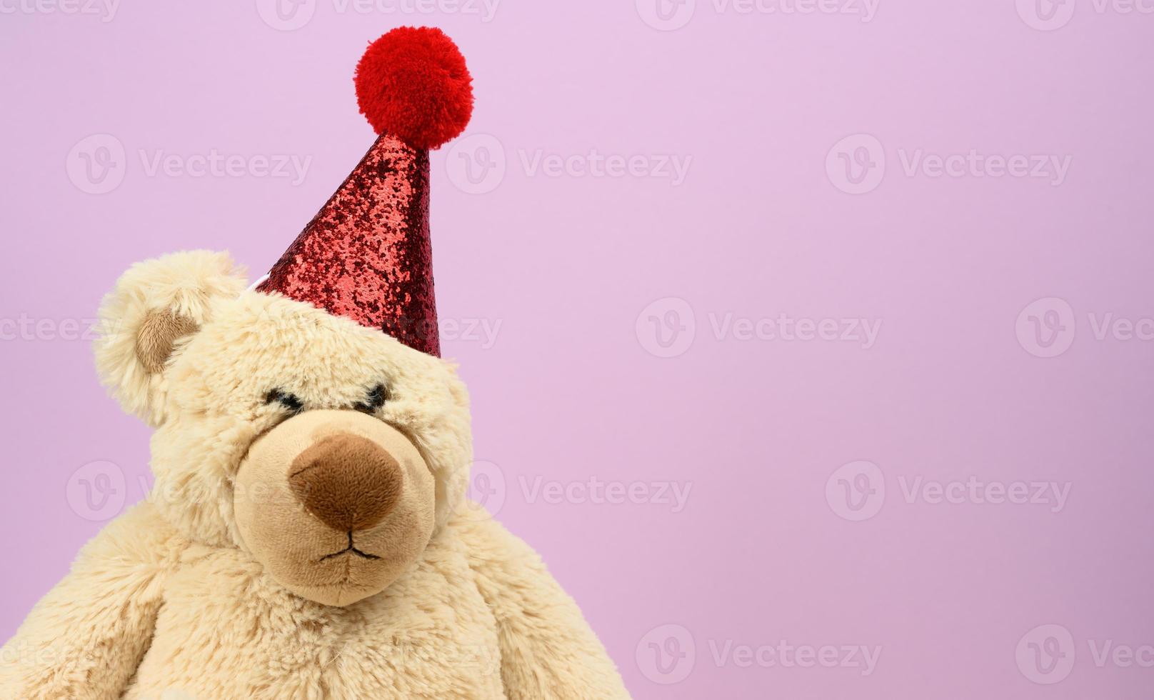 triste ours en peluche beige dans un bonnet rouge est assis sur un fond violet photo