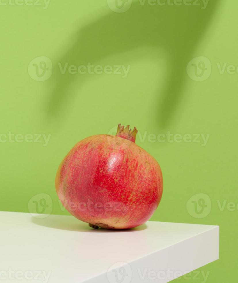 une grenade rouge mûre et entière se trouve sur une table blanche, l'ombre de la main atteint le fruit. fond vert photo