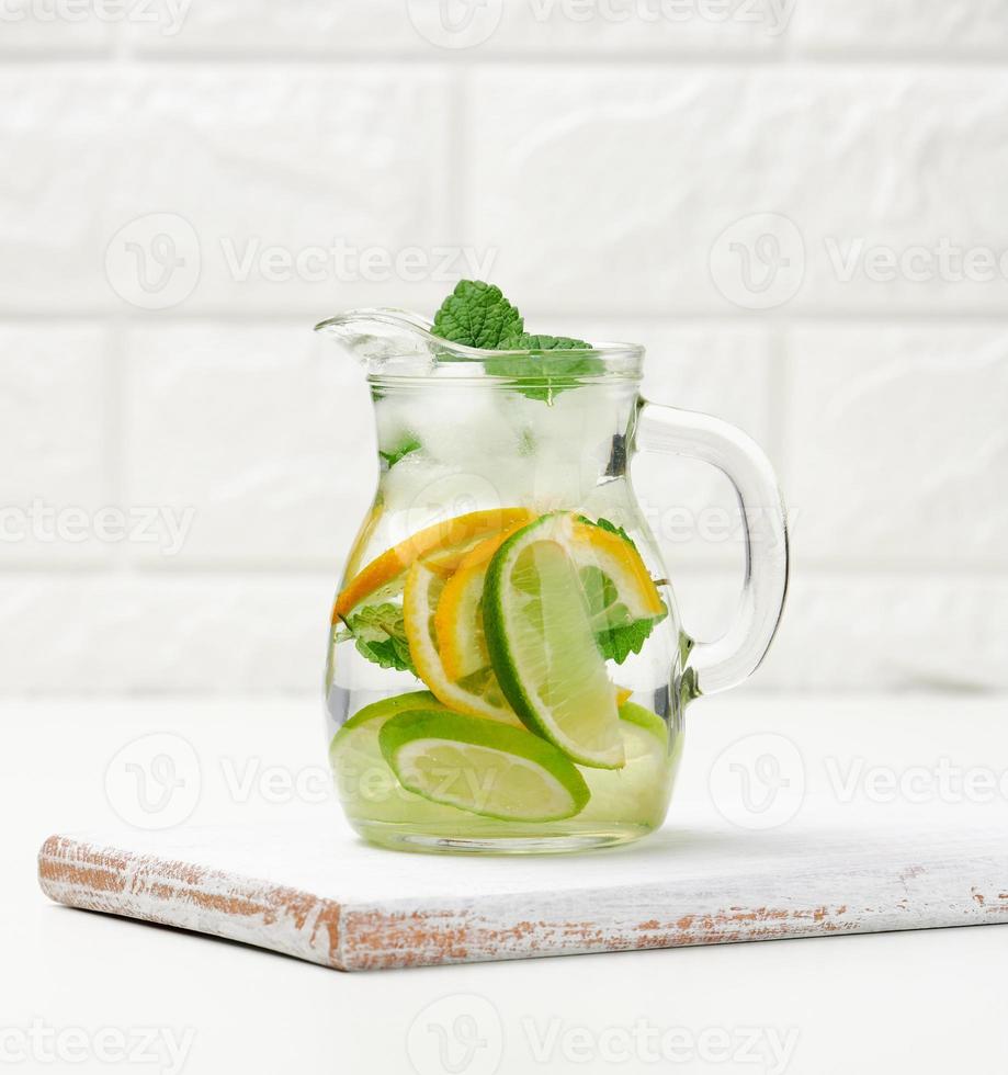 carafe en verre transparent avec tranches de citron, citron vert et feuilles de menthe sur un tableau blanc, désintoxication photo