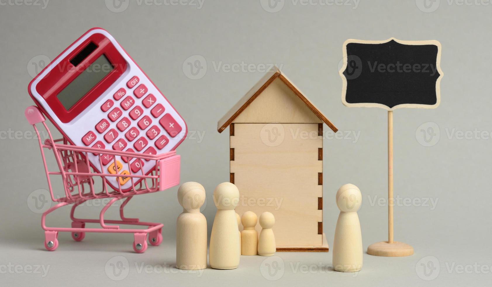 famille de personnages en bois et agent immobilier sur le fond d'une maison et d'une calculatrice dans un chariot sur fond gris photo