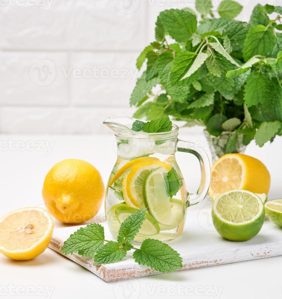 carafe en verre transparent avec tranches de citron, citron vert et feuilles de menthe sur un tableau blanc, détox. derrière les ingrédients de la boisson photo