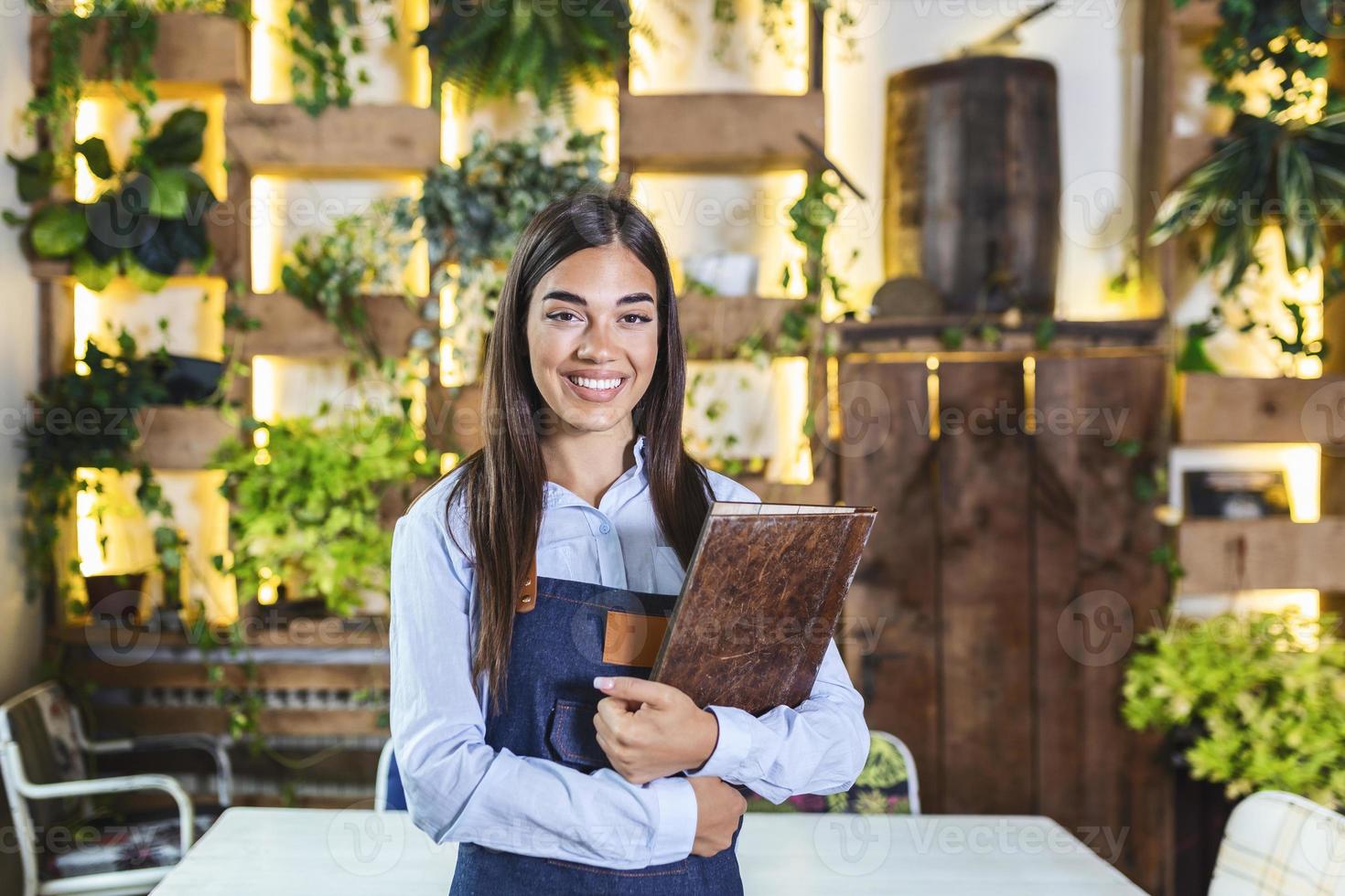 heureuse belle serveuse souriante portant un tablier tenant un menu dossier dans un restaurant, regardant la caméra, debout dans un café confortable, bon service photo