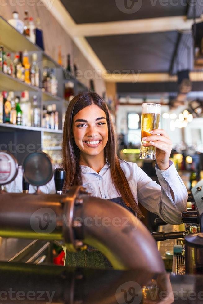 belle barman souriante servant une bière pression au comptoir du bar, jeune femme servant de la bière pression. femme barman souriante versant de la bière fraîche du robinet dans le verre du pub. photo