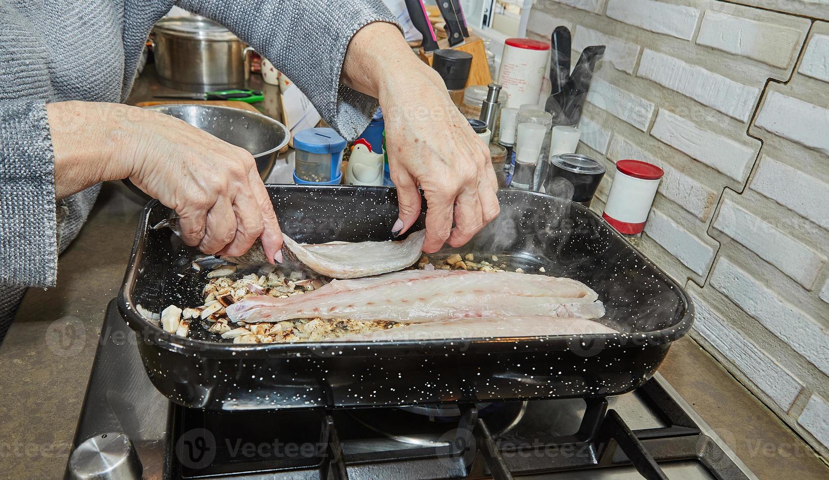 cuisinier met du poisson sur des champignons avec des oignons dans une poêle carrée sur une cuisinière à gaz photo