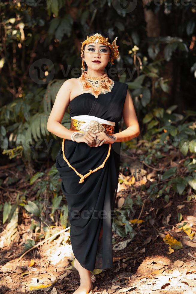 belle femme asiatique debout dans une robe noire tout en portant une couronne dorée et une chaîne dorée à l'intérieur de la forêt photo