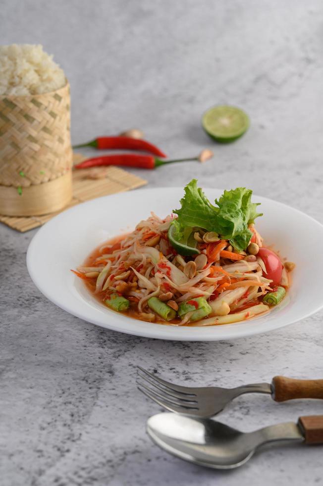 Salade de papaye thaï sur une assiette blanche avec riz gluant, piment, cuillère et fourchette photo