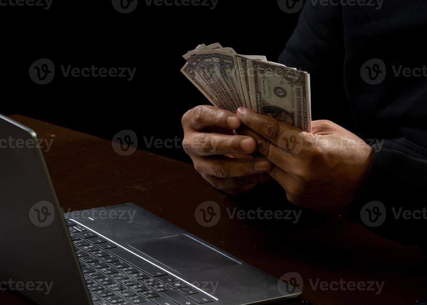 l'ordinateur et les mains d'un homme vêtu d'une chemise noire, assis sur une chaise et une table, est un voleur, détenant de l'argent, comptant le montant obtenu par détournement ou vol, dans une pièce noire. photo