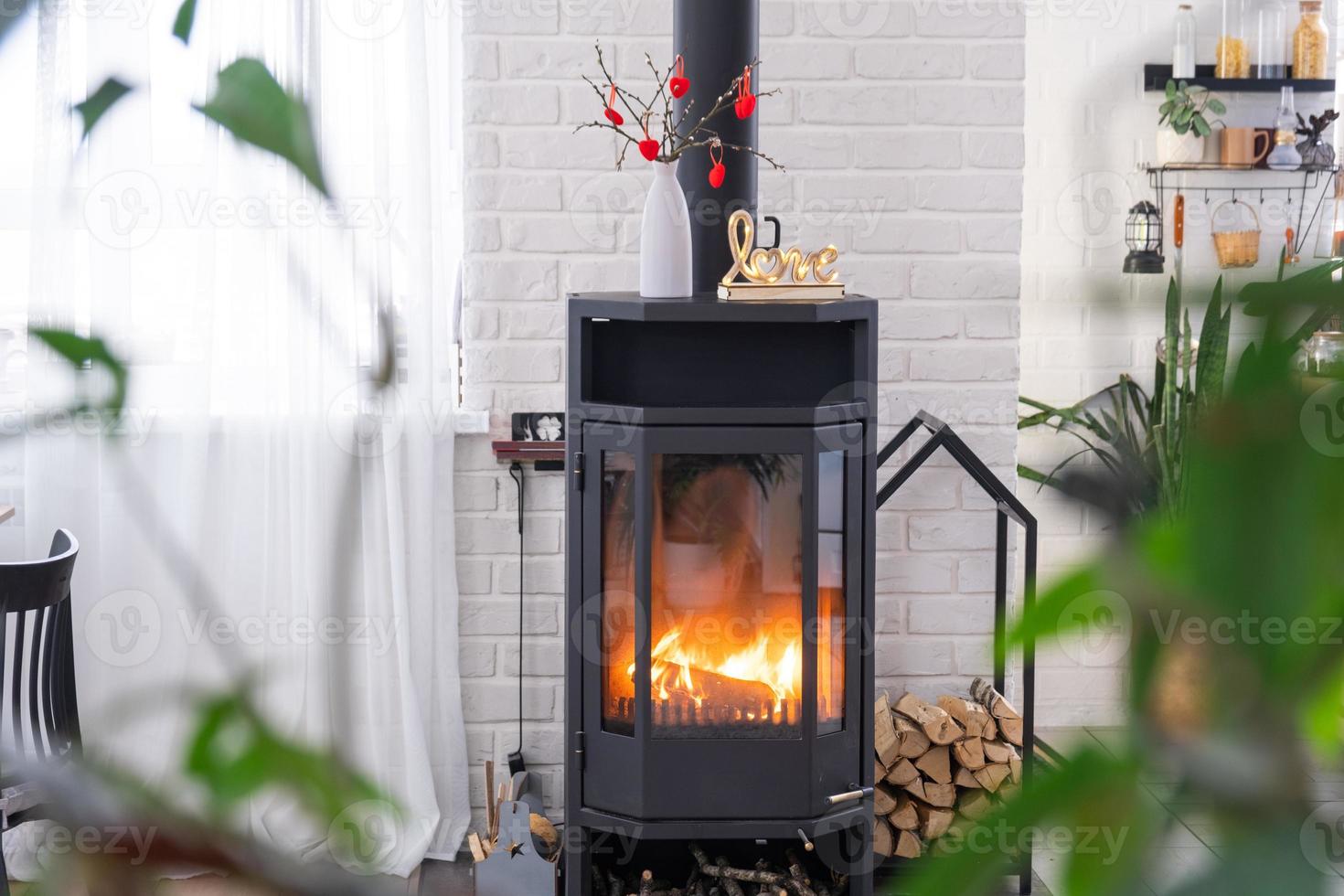 décor de saint valentin près d'un poêle à bois avec feu et bois de chauffage. foyer confortable à l'intérieur avec des plans en pot, saint valentin dans un nid d'amour familial. photo