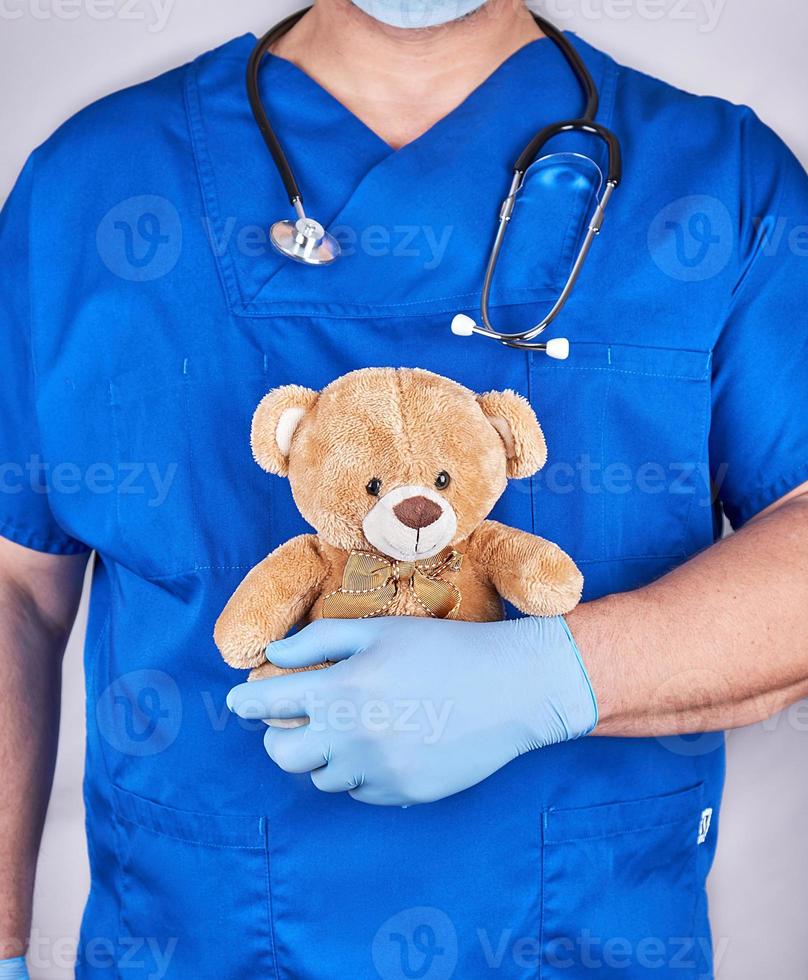 médecin en uniforme bleu et vieux gants en latex tenant un ours en peluche brun photo