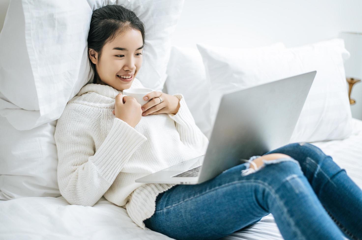 jeune femme jouant sur ordinateur portable et tenant une tasse de café au lit photo