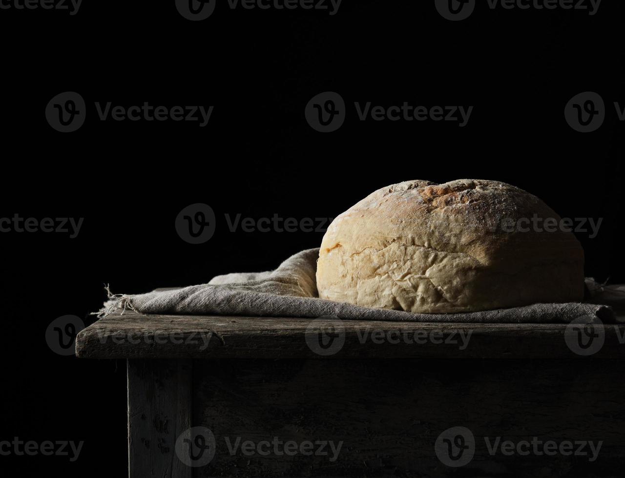 pain de blé blanc rond cuit au four sur une serviette textile, vieille table en bois, fond noir photo