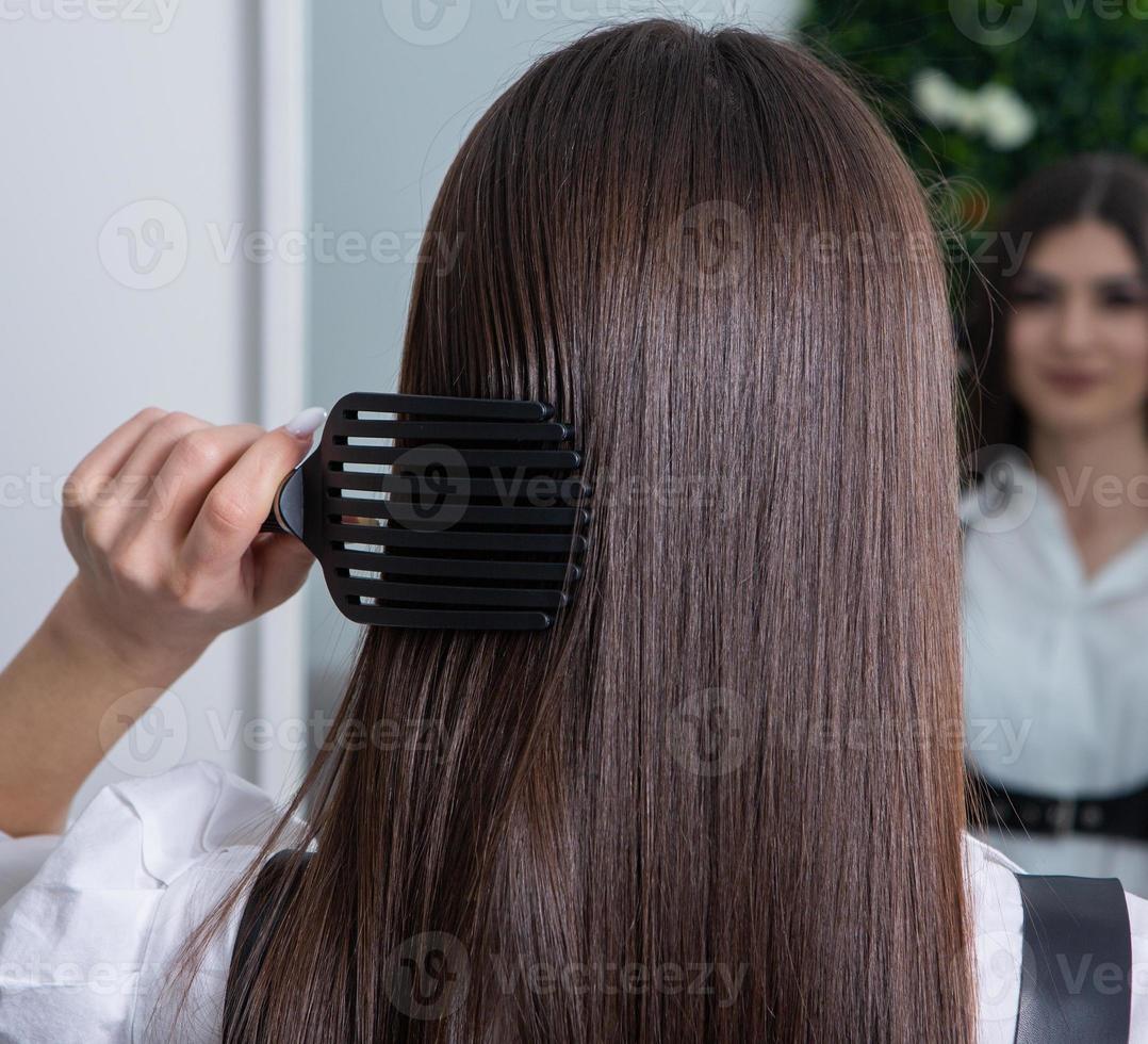 jeune femme peignant ses longs cheveux noirs avec un peigne dans un salon de beauté. une chevelure brune droite et saine qui a subi la procédure de lissage des cheveux. photo