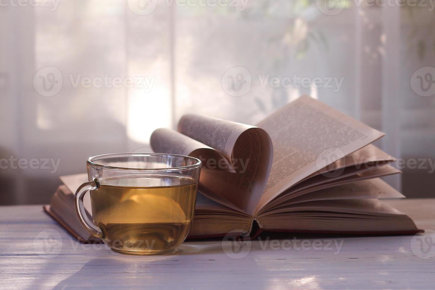 une tasse de thé vert, un livre ouvert sur une table en bois blanche sur fond de fenêtre au soleil. coeur des pages du livre. amour pour la lecture. journée des amoureux des livres photo