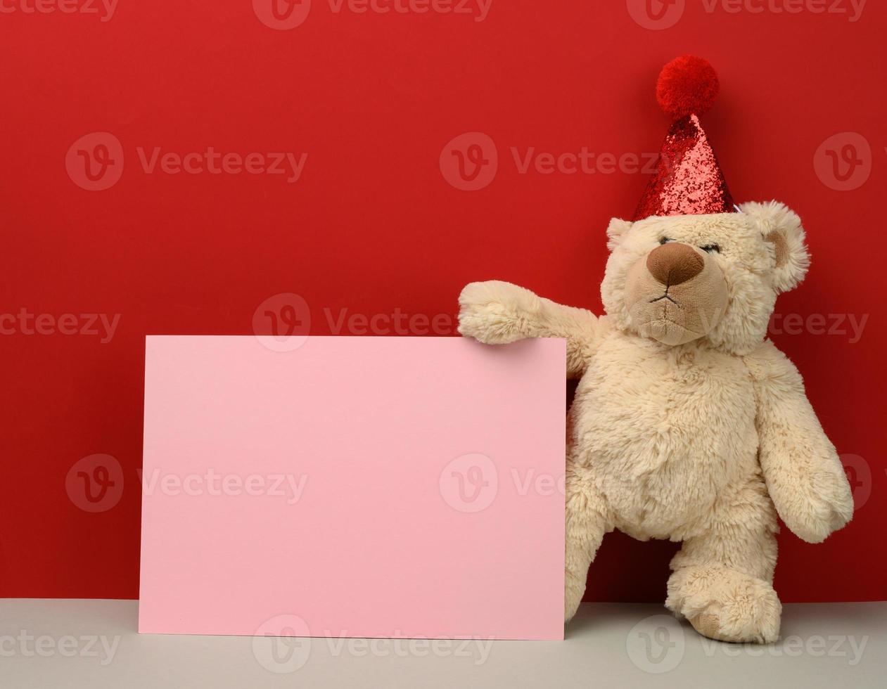 ours en peluche dans un chapeau de fête rouge tient une feuille de papier rose sur fond rouge photo