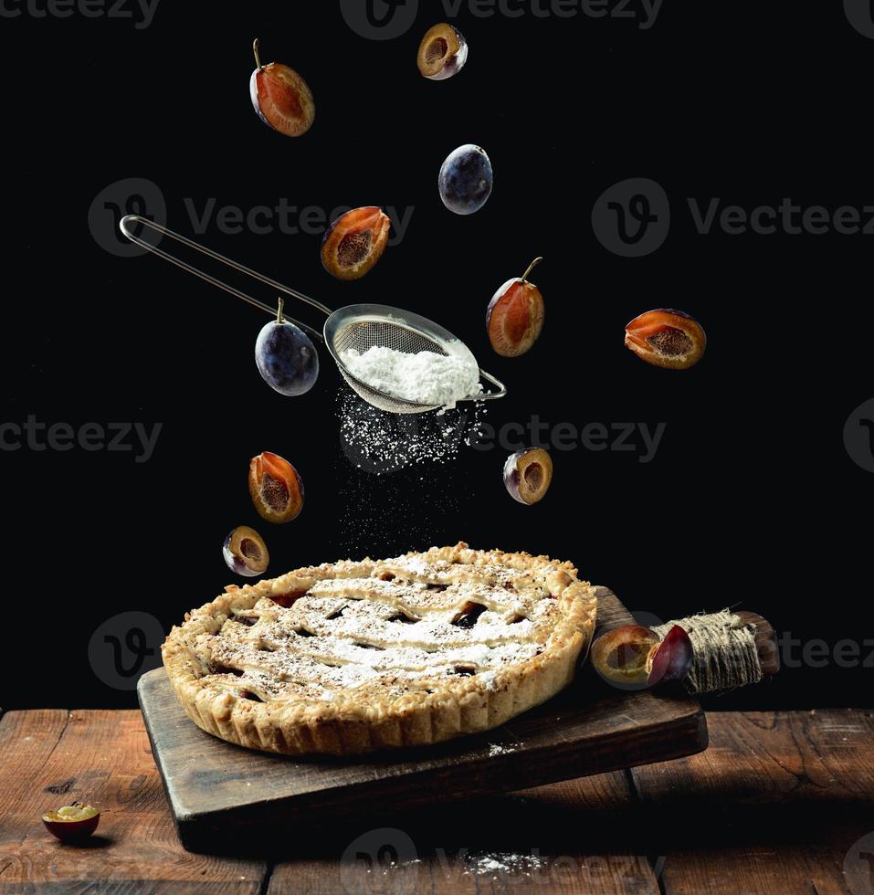 tarte ronde cuite au four avec des prunes sur une planche de cuisine en bois en lévitation, dessus d'un tamis avec du sucre en poudre et des tranches de prunes vivifiantes sur fond noir photo