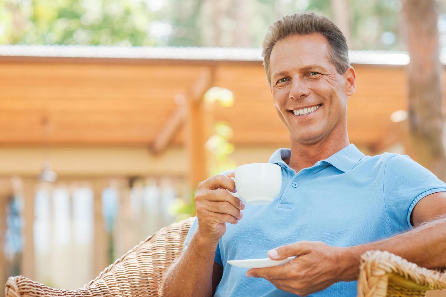 café temps. content mature homme en buvant café et souriant tandis que séance dans chaise en plein air avec maison dans le Contexte photo