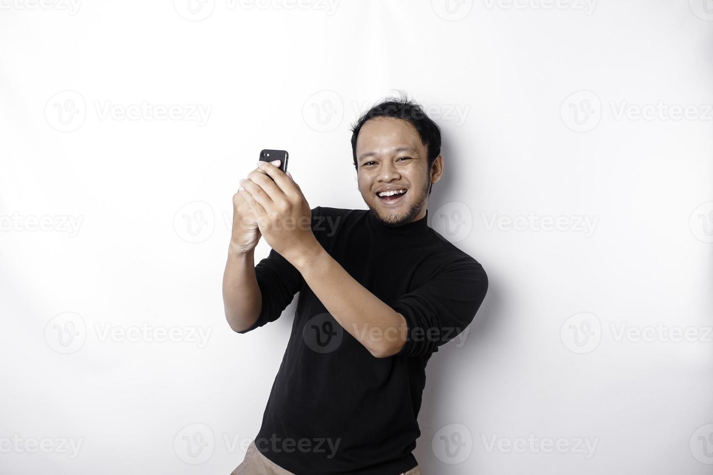 une Jeune asiatique homme avec une content réussi expression portant noir chemise et en portant le sien téléphone, isolé par blanc Contexte photo