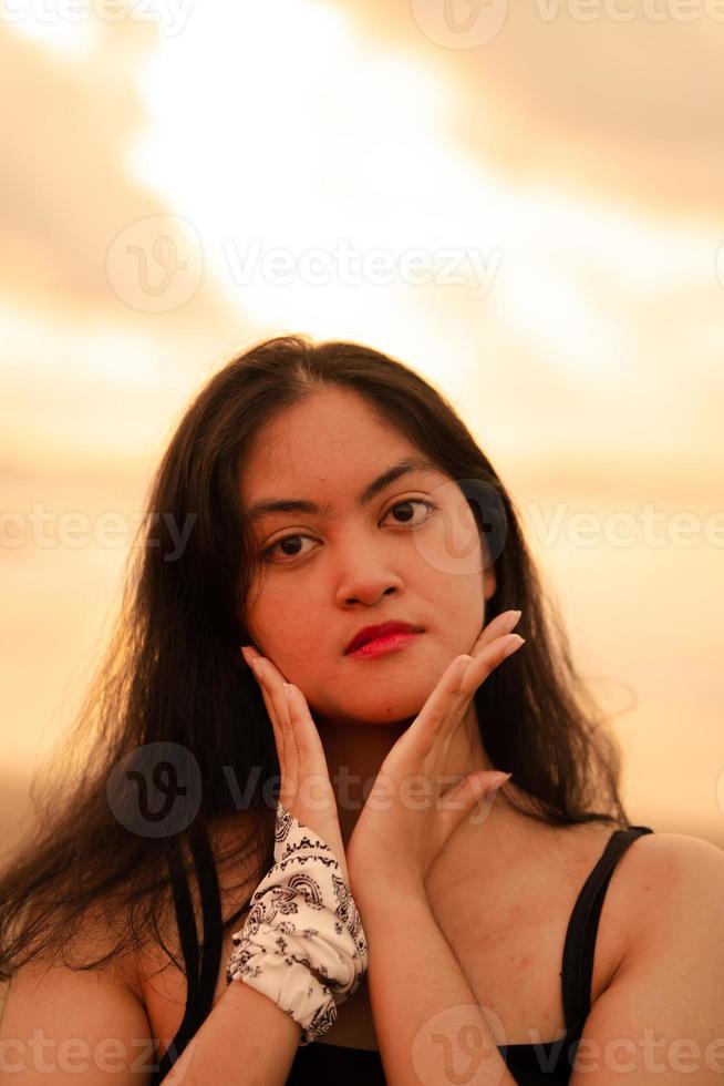 un asiatique femme avec longue noir cheveux pose avec sa mains tandis que profiter le vue de le plage photo