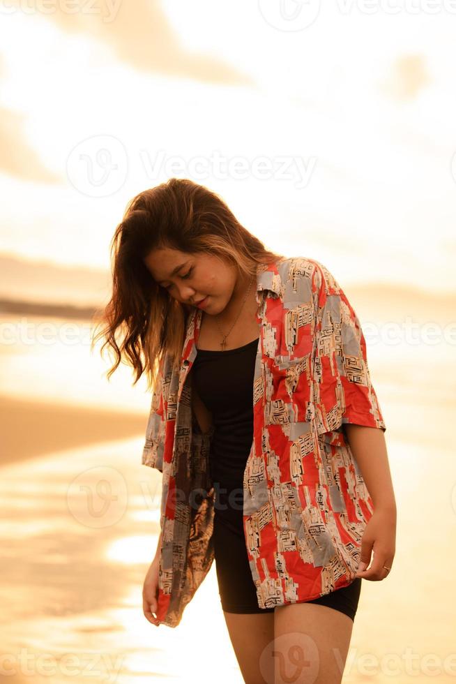 un asiatique femme avec blond cheveux et une rouge chemise des promenades le long de le plage à prendre plaisir le mer vue photo