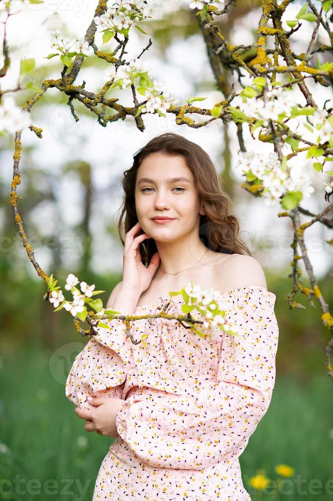 une fille dans une rose robe des stands près une blanc arbre avec fleurs regards à le caméra et sourit photo