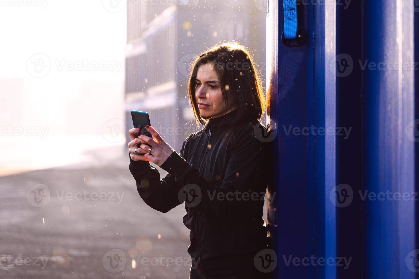 Jeune adulte femelle coureur est en utilisant téléphone intelligent parmi le conteneurs de une commercial Port photo