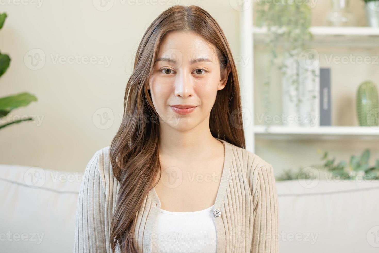 souriant positif, attrayant asiatique Jeune femme portant décontractée robe, portrait de magnifique brunette sa avec longue marron cheveux, sentiment content à la recherche à caméra séance sur canapé, isolé sur Contexte. photo