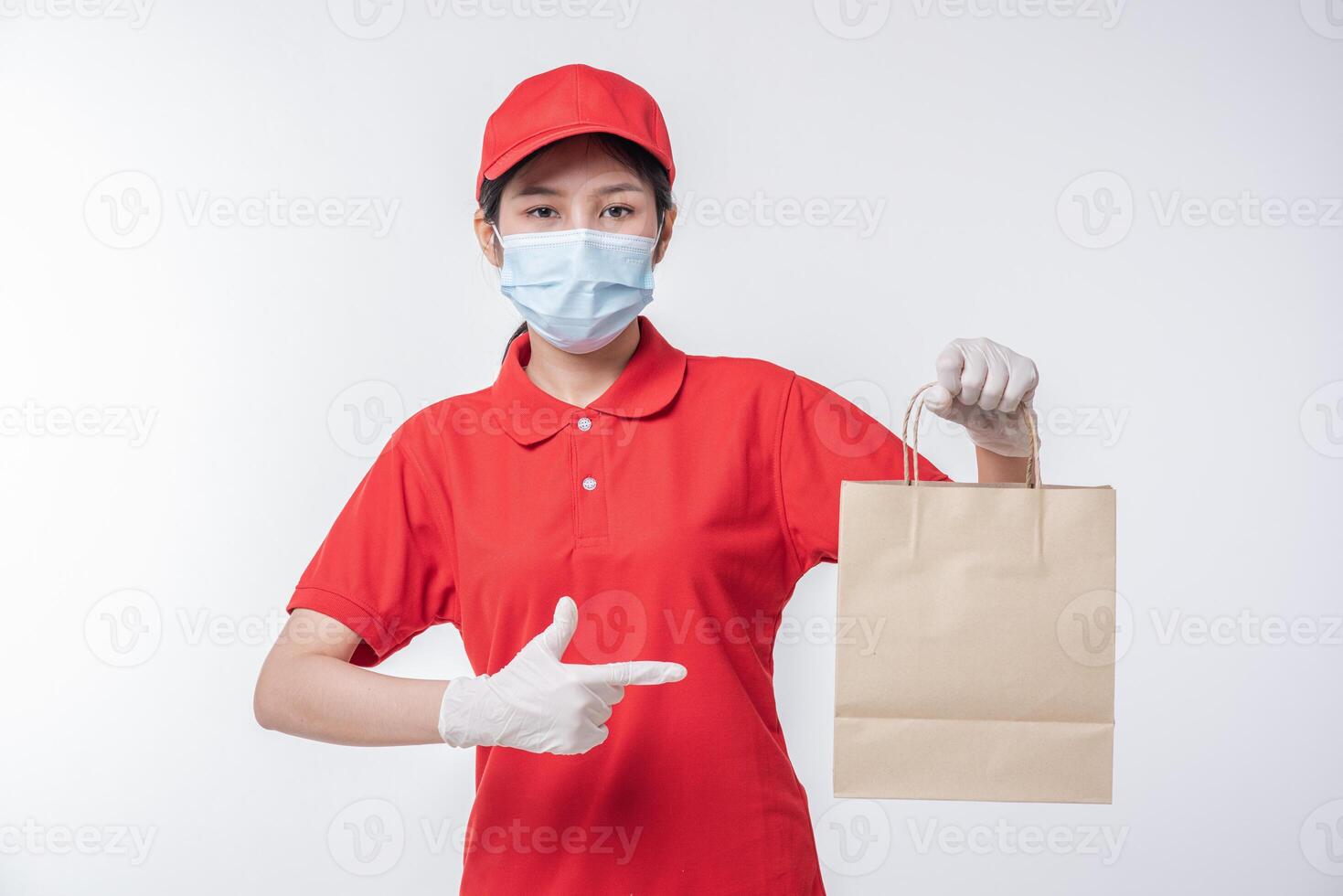 image d'un jeune livreur heureux en casquette rouge t-shirt blanc uniforme masque facial gants debout avec paquet de papier kraft marron vide isolé sur fond gris clair studio photo