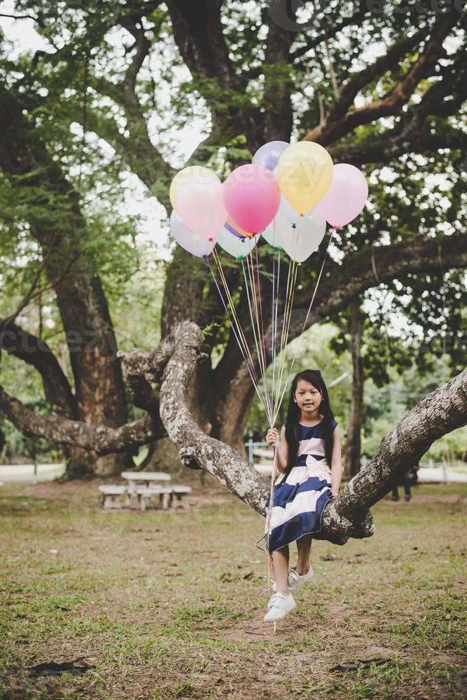 petite fille asiatique mignonne assise sur une branche d'arbre avec des ballons colorés photo