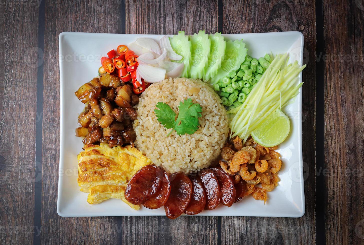 Riz assaisonné de pâte de crevettes, avec oignons rouges, haricots, mangue et œuf au plat sur plaque blanche sur table en bois photo