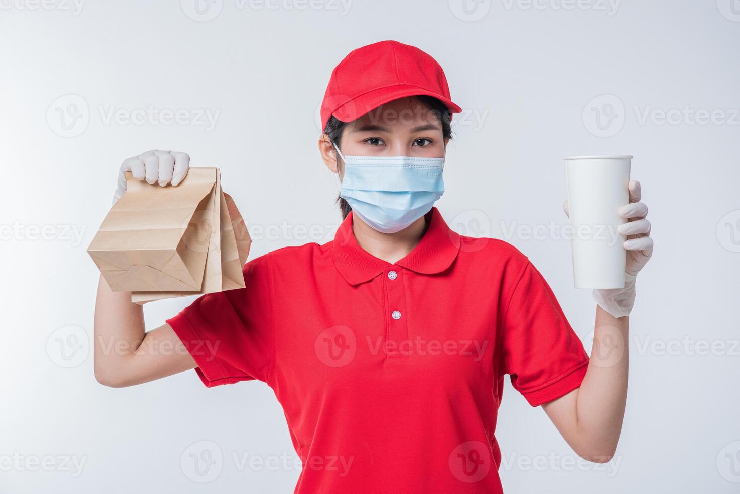 image d'un jeune livreur heureux en casquette rouge t-shirt blanc uniforme masque facial gants debout avec paquet de papier kraft marron vide isolé sur fond gris clair studio photo