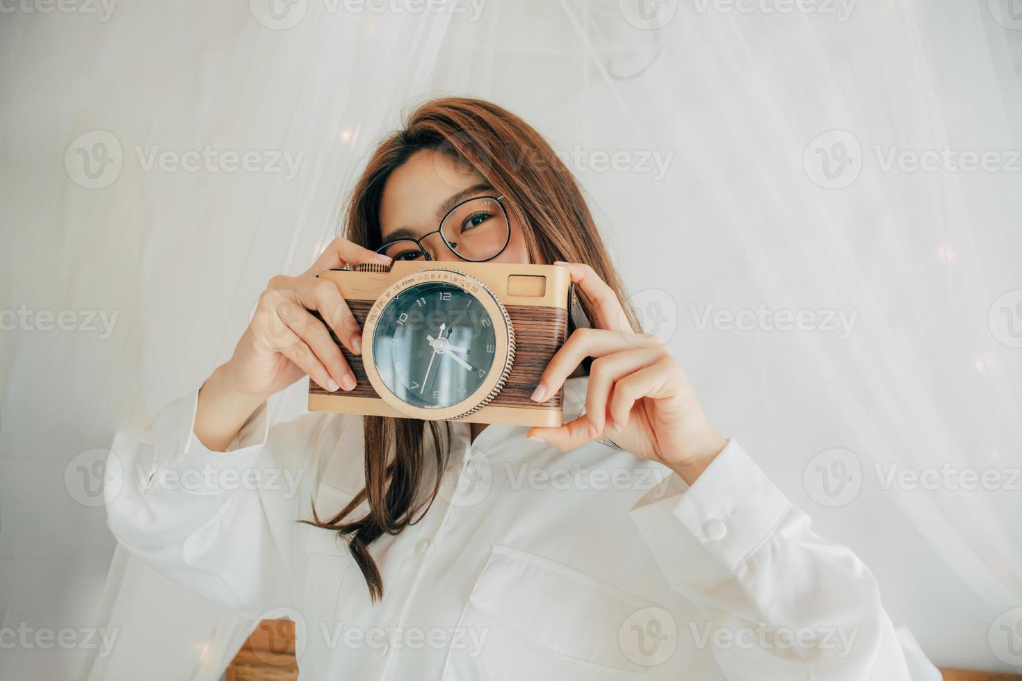 portrait d'une belle femme asiatique heureuse souriante en chemise blanche posant sur le lit. prendre des photos à partir d'une horloge qui ressemble à un appareil photo. concept de mode de vie des gens. espace de copie. regardant la caméra.