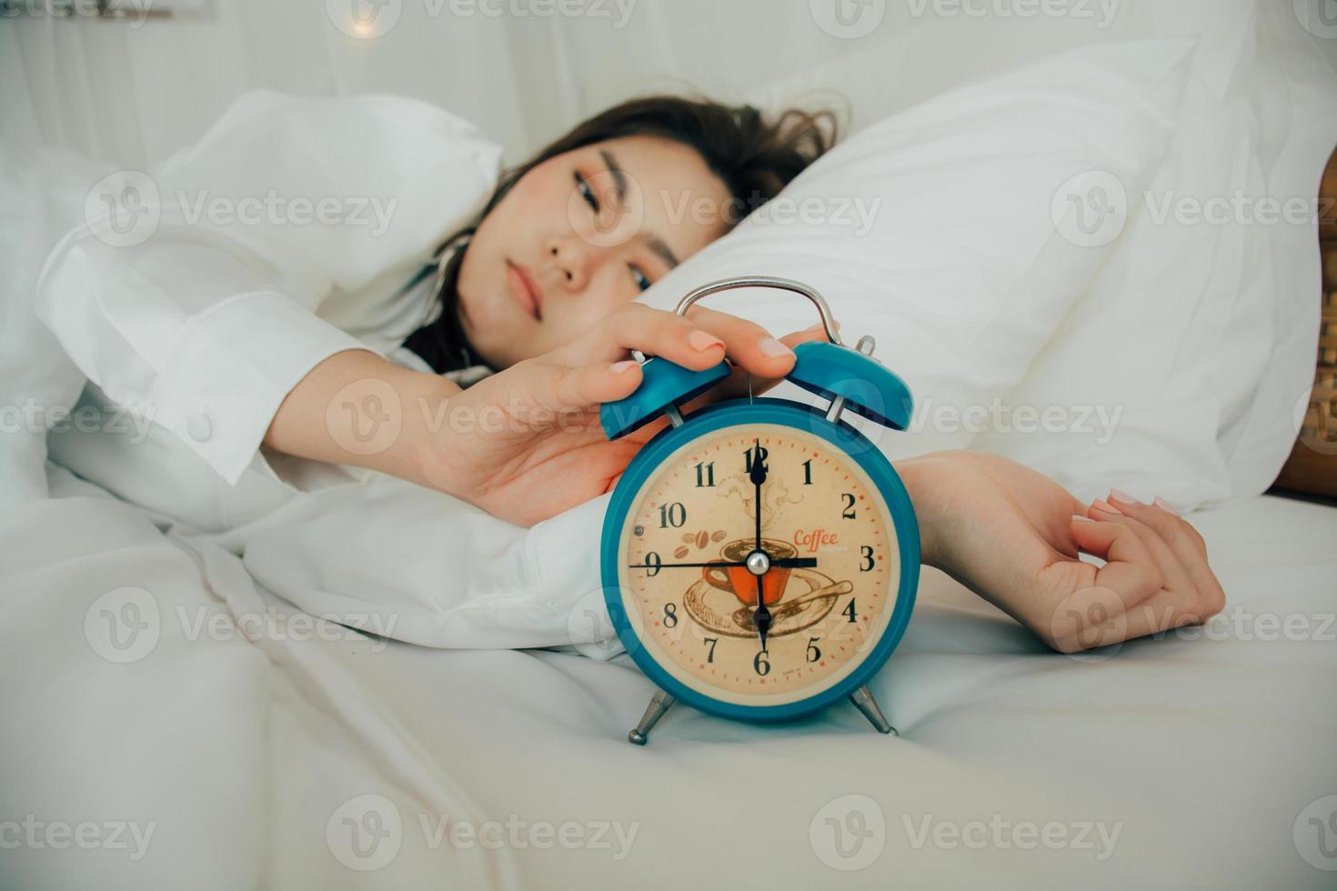 asiatique magnifique femme dans blanc pyjamas tournant de alarme l'horloge tandis que somnolent dans lit à sa chambre de maison sur vacances. fille est ayant difficulté réveiller en haut en retard dans Matin. mauvais pour la santé dormir. sélectif concentrer photo