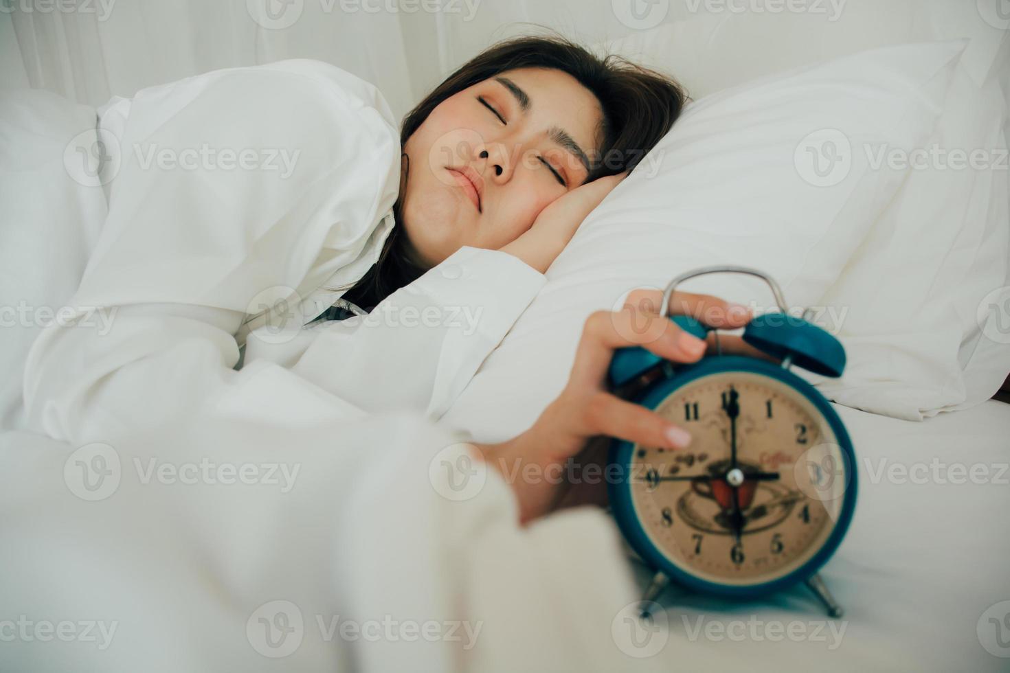 asiatique magnifique femme dans blanc pyjamas tournant de alarme l'horloge tandis que somnolent dans lit à sa chambre de maison sur vacances. fille est ayant difficulté réveiller en haut en retard dans Matin. mauvais pour la santé dormir. sélectif concentrer photo