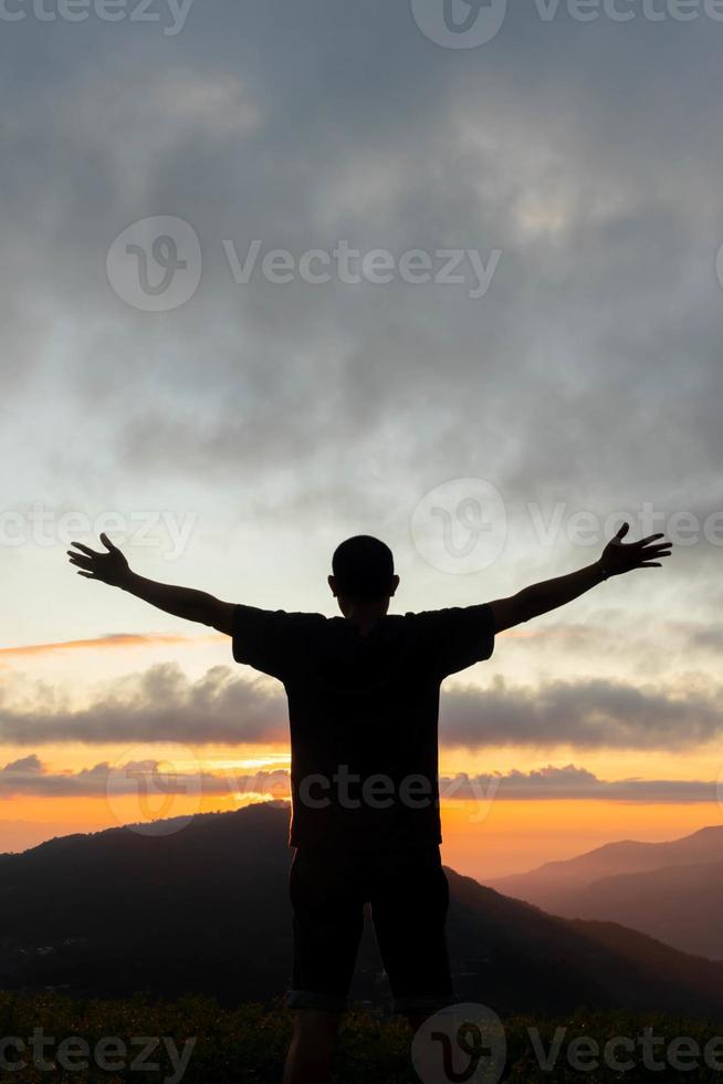 silhouette de Jeune homme permanent sur Haut de Montagne de pointe avec le sien mains élevé au dessus le sien tête comme symbole de culte et invocation de dieux bénédictions avec croyance et Puissance de Foi dans Dieu. photo