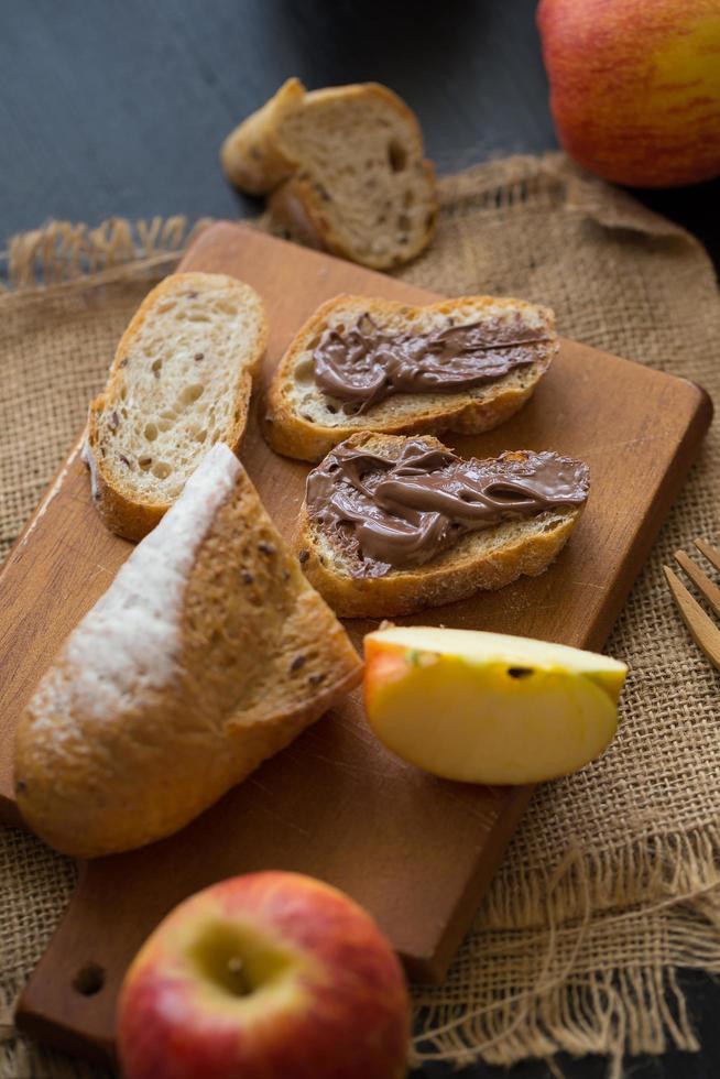 Pommes entières et tranchées avec baguette en tranches avec un beurre au chocolat sur planche de bois sur une table en bois foncé photo