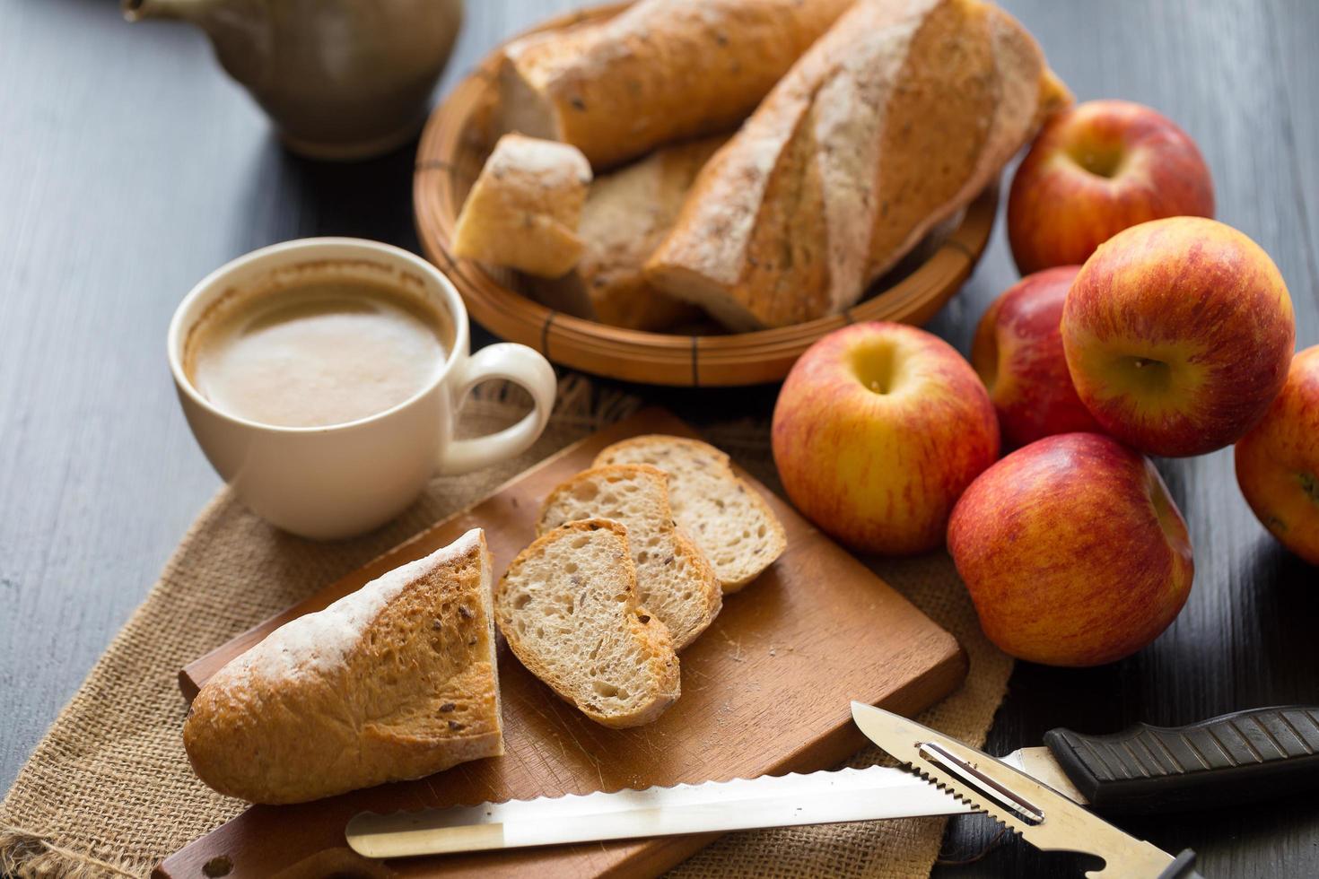 Pommes entières avec baguette en tranches sur planche de bois avec des couteaux et une tasse de café sur une table en bois sombre photo